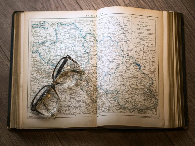 A close-up of a vintage map with eyeglasses resting on an open book, set on a wooden table.